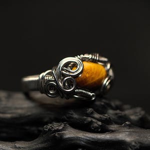 Könnte beinhalten: Ein Ring aus silbernem Draht, umwickelt mit einem Tigerauge-Edelstein. Der Ring liegt auf einer dunklen Holzoberfläche.