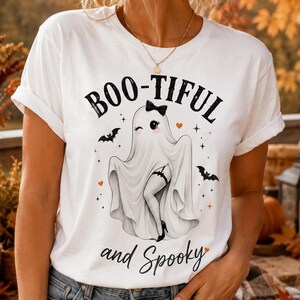 Peut inclure: Un t-shirt blanc à manches courtes avec un motif de fantôme portant un nœud noir et des bas résille. Le texte indique BOO-TIFUL and Spooky, accompagné de petites chauves-souris noires, de cœurs orange et d'étoiles imprimés sur le devant.
