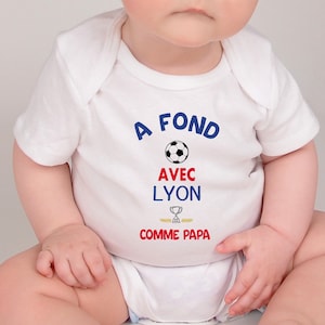 Peut inclure: Body bébé blanc avec l'inscription "A FOND AVEC LYON COMME PAPA" en bleu et rouge, avec un ballon de foot et un trophée. Le body est conçu pour les nourrissons.