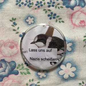 Può includere: Un bottone rotondo con un'illustrazione di uccello e il testo tedesco "Lass uns auf Nazis scheißen." Il bottone ha una copertura trasparente, simile alla plastica, e si trova su uno sfondo di tessuto floreale.