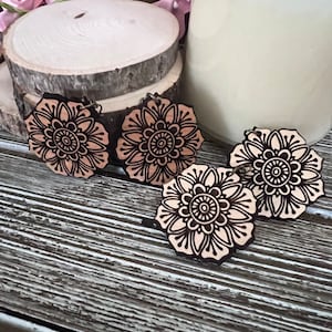 May include: Four wooden flower earrings with a detailed floral design. The earrings are light brown with dark brown etched outlines. The earrings are arranged on a rustic wooden surface, with a candle and decorative wood slices in the background.