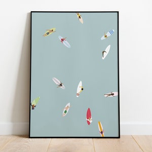 Puede incluir: Una impresión artística enmarcada que presenta un fondo azul claro con múltiples surfistas en tablas de surf. Los surfistas se representan desde una perspectiva cenital, con varios colores y diseños en sus tablas y trajes de baño. La obra está enmarcada en negro.