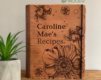 Puede incluir: Un libro de recetas de madera con un diseño floral y el texto "Caroline Mae's Recipes". El libro está cerrado y tiene una banda elástica marrón.