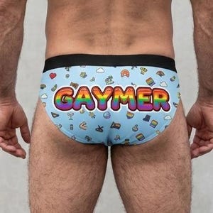 Puede incluir: Calzoncillos azul claro con cintura negra, con un estampado de pequeños iconos y la palabra "GAYMER" en colores arcoíris. Los calzoncillos se muestran por delante y por detrás.