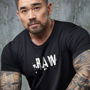 May include: A man with short black hair and a beard is wearing a black t-shirt with the word "RAW" printed on it in white. He has tattoos on both arms.