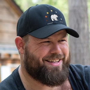 May include: A man with a thick beard is smiling and wearing a black baseball cap with a white bear and stars design. He is wearing a black shirt.