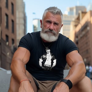 May include: A man with a gray beard and gray hair is wearing a black t-shirt with a black and white graphic of a man in a leather jacket. He is sitting on a concrete surface.