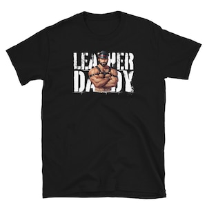 May include: Black t-shirt with white text that reads "Leather Daddy". A man wearing a leather harness and cap is pictured in the center of the shirt.
