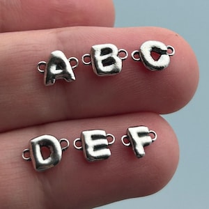 Puede incluir: Seis pequeños dijes plateados con letras del alfabeto: A, B, C, D, E y F. Cada dije tiene un lazo a cada lado para sujetarlo a una pulsera o collar. Los dijes se muestran sobre un fondo azul claro.