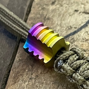 May include: A multi-colored metal bead with a ridged design is attached to a green paracord.