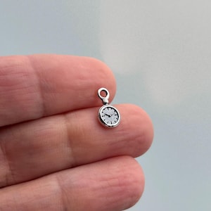 May include: A small, silver-colored charm in the shape of a pocket watch. The watch face is white with black hands and hour markers. The charm has a small loop at the top for attaching to a bracelet or necklace.
