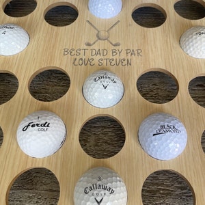 May include: A wooden golf ball display with multiple golf balls in circular cutouts. The display has the text "BEST DAD BY PAR LOVE STEVEN" and a crossed golf club graphic. The golf balls are white with various brand names.