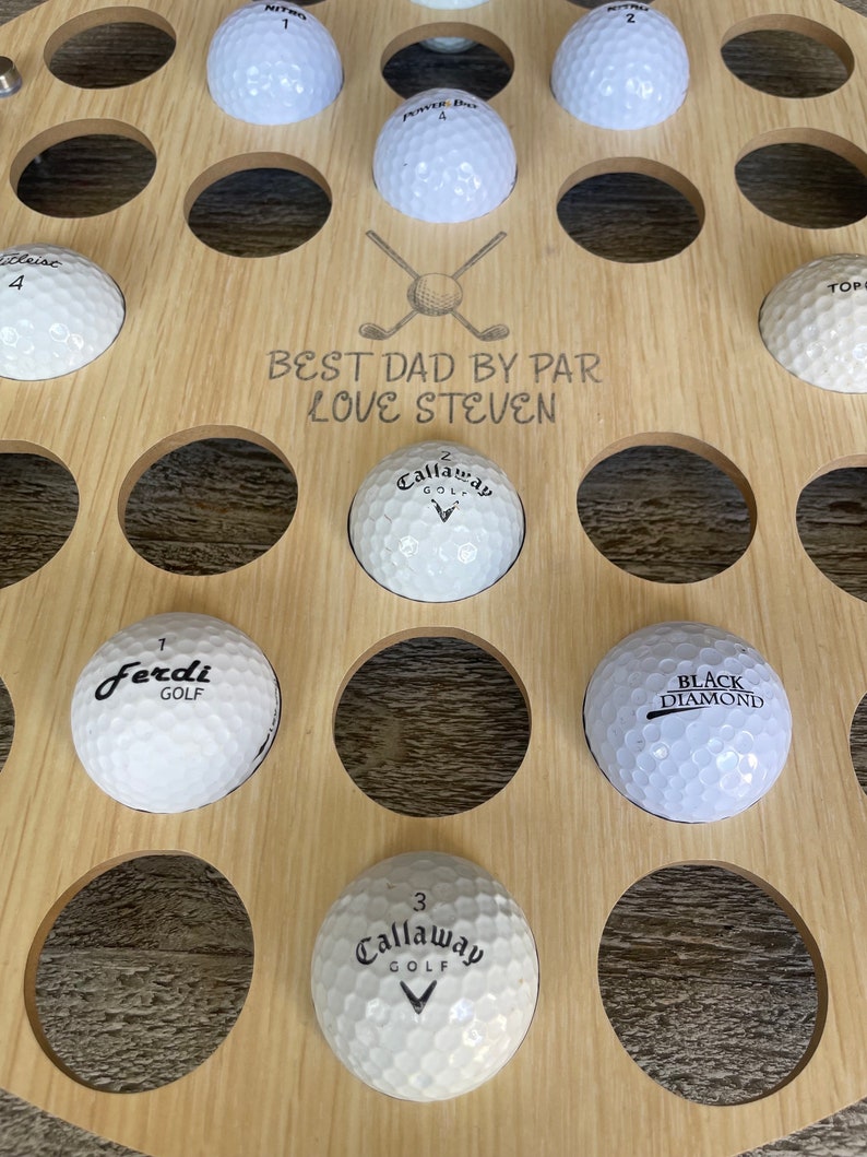 May include: A wooden golf ball display with multiple golf balls in circular cutouts. The display has the text "BEST DAD BY PAR LOVE STEVEN" and a crossed golf club graphic. The golf balls are white with various brand names.