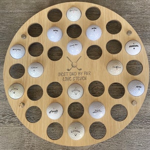 May include: A round, wooden golf ball display with 28 holes, holding white golf balls. The display has the text "BEST DAD BY PAR LOVE STEVEN" and crossed golf clubs. Metal studs secure the display. Golf balls are labeled with brand names and numbers.