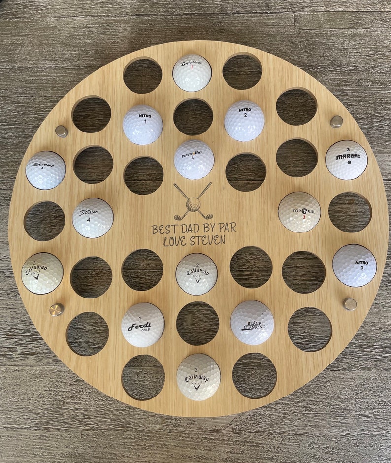 May include: A round, wooden golf ball display with 28 holes, holding white golf balls. The display has the text "BEST DAD BY PAR LOVE STEVEN" and crossed golf clubs. Metal studs secure the display. Golf balls are labeled with brand names and numbers.