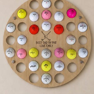 May include: A wooden golf ball display with 30 holes, each holding a golf ball of various colors. The display has the text "BEST DAD BY PAR LOVE EMILY" and is mounted on a light-colored wall.