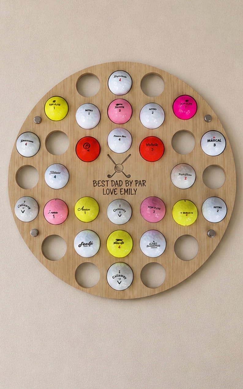 May include: A wooden golf ball display with 30 holes, each holding a golf ball of various colors. The display has the text "BEST DAD BY PAR LOVE EMILY" and is mounted on a light-colored wall.