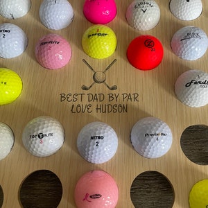 May include: A wooden golf ball display with multiple golf balls in various colors, including pink, yellow, and white. The display has the text "BEST DAD BY PAR LOVE HUDSON" and a golf club graphic.