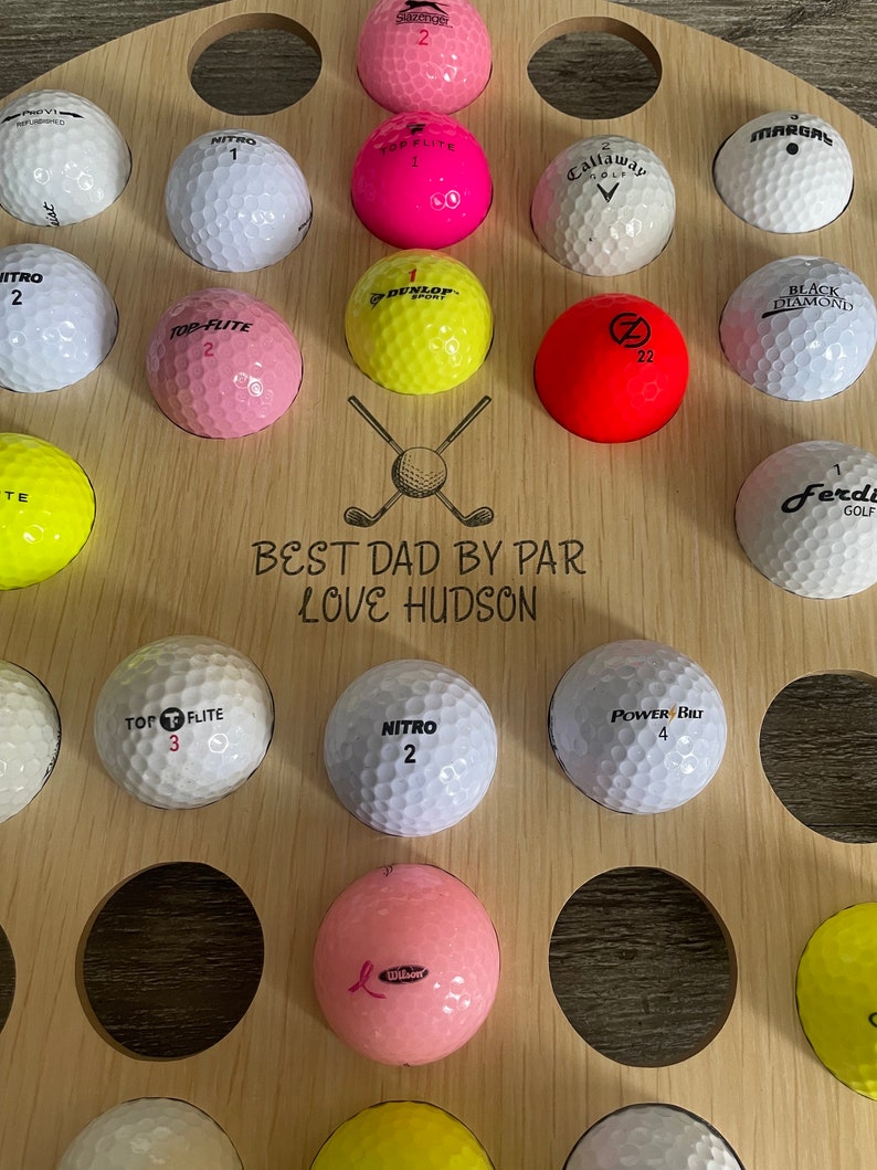 May include: A wooden golf ball display with multiple golf balls in various colors, including pink, yellow, and white. The display has the text "BEST DAD BY PAR LOVE HUDSON" and a golf club graphic.