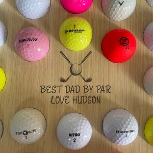 May include: Wooden golf ball display board with various colored golf balls. The board has the text "BEST DAD BY PAR LOVE HUDSON" and a graphic of crossed golf clubs and a golf ball. Golf balls are white, pink, yellow, and red.