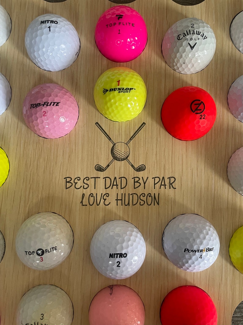 May include: Wooden golf ball display board with various colored golf balls. The board has the text "BEST DAD BY PAR LOVE HUDSON" and a graphic of crossed golf clubs and a golf ball. Golf balls are white, pink, yellow, and red.