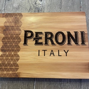 May include: A wooden cutting board with a geometric pattern on the left side and the words "PERONI ITALY" engraved in black on the right side.
