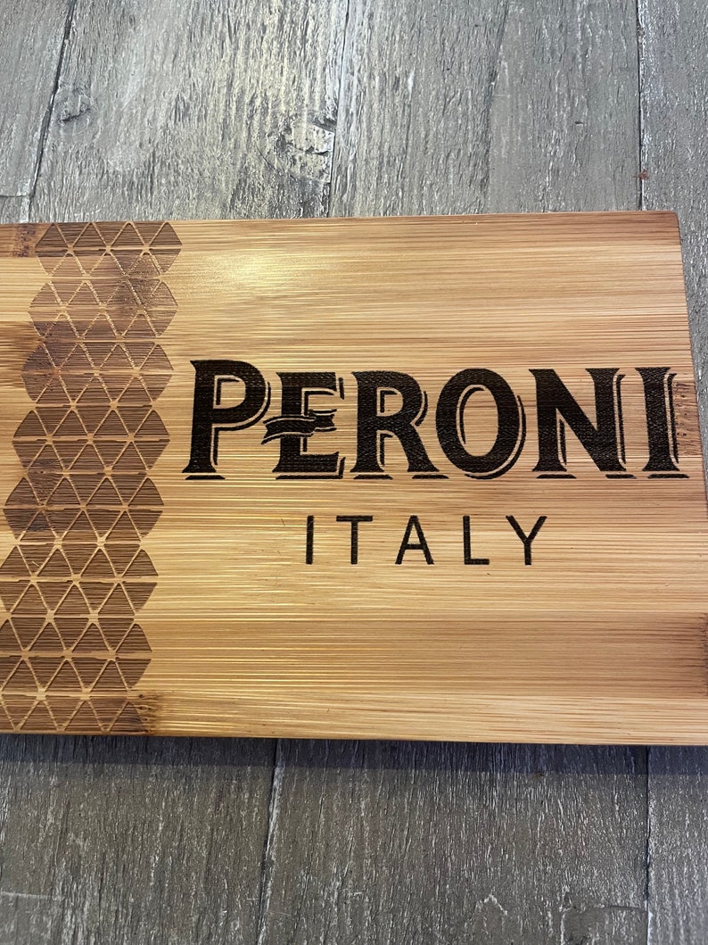 May include: A wooden cutting board with a geometric pattern on the left side and the words "PERONI ITALY" engraved in black on the right side.