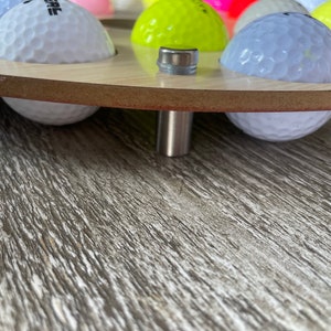 May include: A wooden golf ball display stand with multiple golf balls in various colors. The stand has a light wood finish and a metal support. The golf balls are white, yellow, pink, and blue.