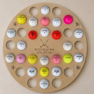 May include: A round wooden golf ball display with 28 golf balls of various colors and brands. The display has a circular design with holes to hold the balls. Text on the display reads "BEST DAD BY PAR LOVE STEVEN."