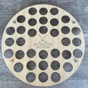 May include: A round, light-colored wooden golf ball display with numerous circular holes. The text "BEST DAD BY PAR LOVE STEVEN" is engraved below a golf ball and crossed club graphic. Metal fasteners secure the display.