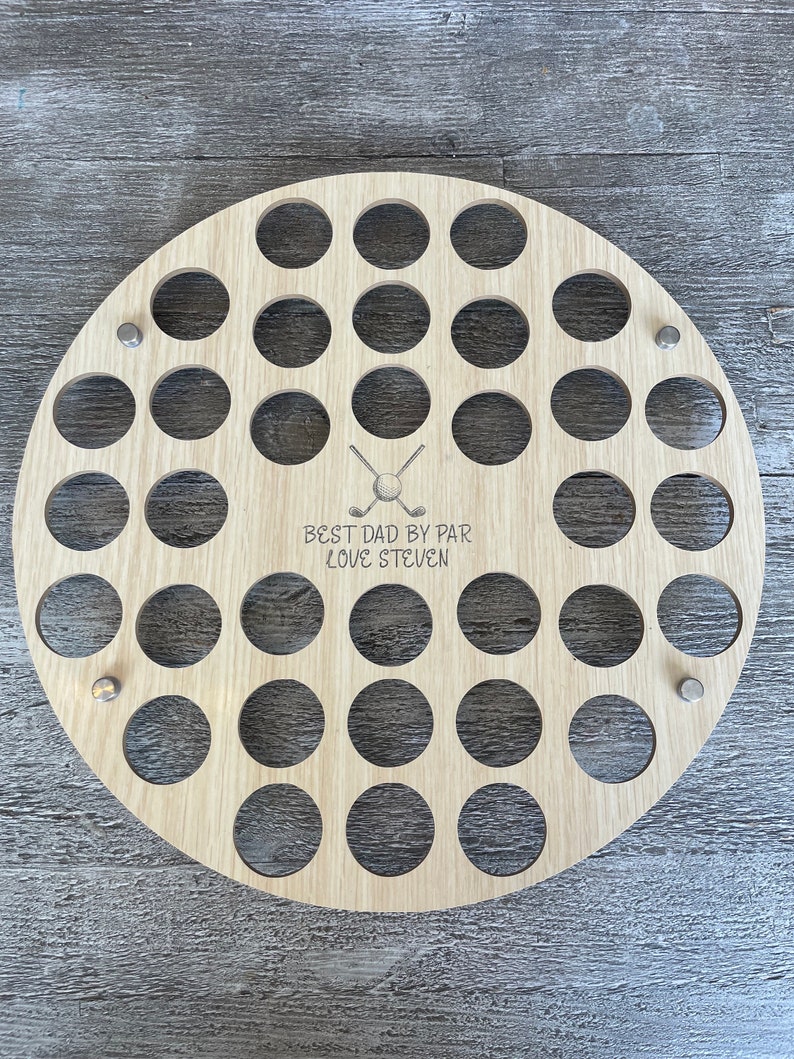 May include: A round, light-colored wooden golf ball display with numerous circular holes. The text "BEST DAD BY PAR LOVE STEVEN" is engraved below a golf ball and crossed club graphic. Metal fasteners secure the display.