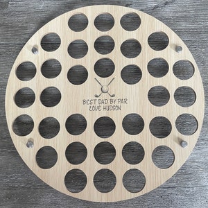 May include: A round, light-colored wooden golf ball display with 30 circular holes. The display has the text "BEST DAD BY PAR LOVE HUDSON" and a crossed golf club and ball graphic. Three silver pins are used to mount the display.