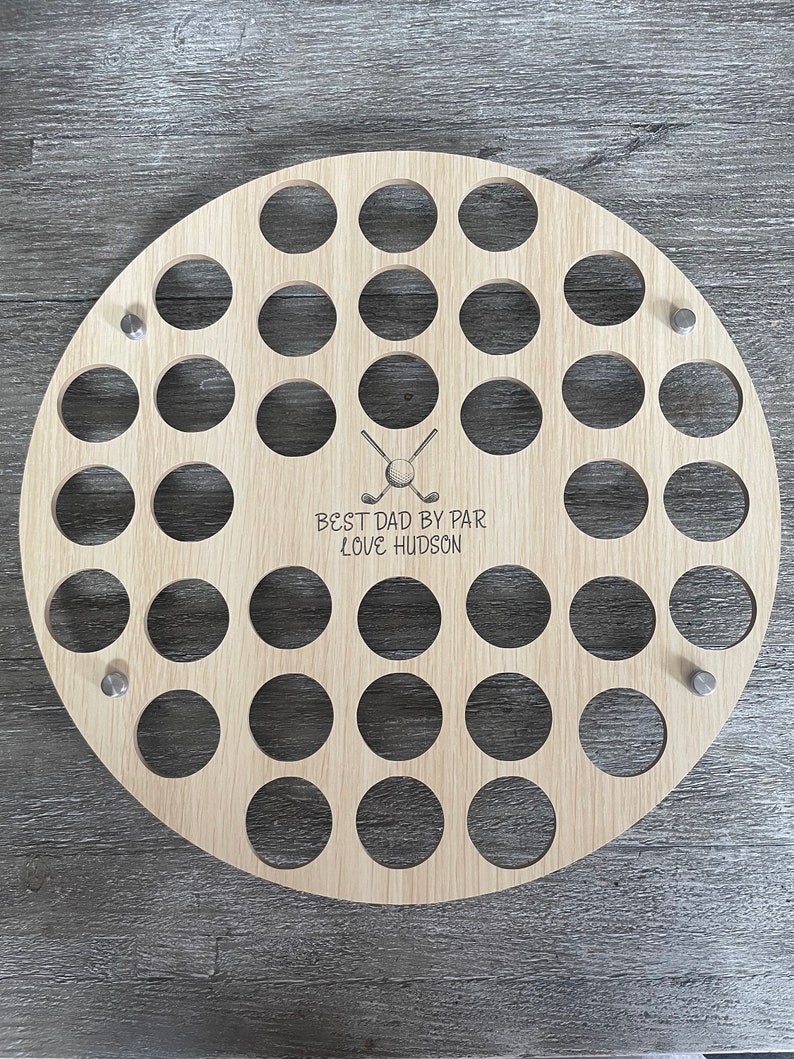 May include: A round, light-colored wooden golf ball display with 30 circular holes. The display has the text "BEST DAD BY PAR LOVE HUDSON" and a crossed golf club and ball graphic. Three silver pins are used to mount the display.