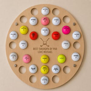 May include: A round wooden golf ball display board with holes holding various golf balls. The balls are different colors and brands. The text "BEST GRANDPA BY PAR LOVE MICHAEL" is in the center.