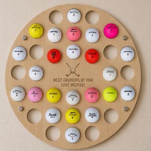 May include: A round wooden golf ball display with 25 golf balls of various brands and colors. The display has a personalized message that reads "BEST GRANDPA BY PAR LOVE MICHAEL".