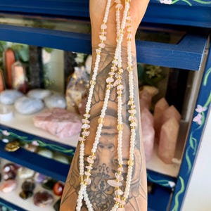 May include: Three strands of beaded necklaces with white beads, pink and yellow gemstones, and gold-colored accents. The necklaces are held up against a blue cabinet with floral designs and a display of crystals.