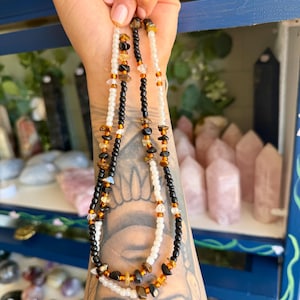 May include: A multi-strand beaded necklace featuring a combination of small white, black, and translucent beads interspersed with irregular, polished brown and amber-colored gemstone chips, creating a layered and textured appearance.