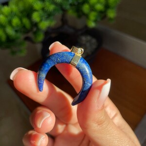 May include: A crescent-shaped pendant made of deep blue lapis lazuli, with gold-colored metal accents. The gemstone has a speckled appearance. The pendant is held up by a hand, with a blurred green plant in the background.