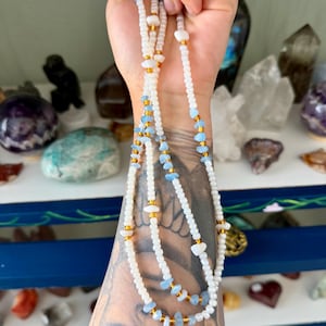May include: A long beaded necklace featuring a repeating pattern of small white seed beads, metallic gold spacer beads, and irregular light blue and white stone chips. The necklace is strung on a flexible cord and hangs in multiple loops.