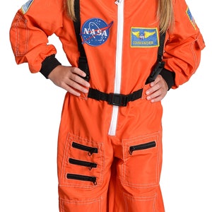 May include: A child wearing an orange astronaut costume with NASA and Commander patches. The costume has a black belt, white stitching, and multiple pockets with zips. The child is also wearing a black baseball cap with a white NASA logo.