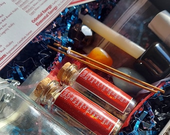 May include: A box filled with various items for a spiritual ritual, including two small glass bottles with cork tops labeled "Health Herbs", a white candle, a small bag, a clear glass bottle, a small orange crystal, and two incense sticks. The box is lined with blue and black shredded paper.