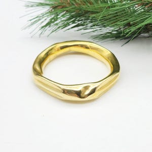 May include: A gold-coloured, irregularly shaped bangle bracelet. The bracelet has a smooth, polished surface and a slightly uneven texture. The background is white with a sprig of green foliage.