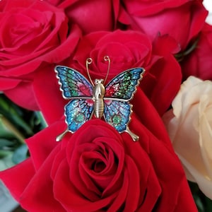 Puede incluir: Un broche de mariposa colorido con detalles azules, verdes y rojos descansa sobre una rosa roja vibrante. El broche tiene un cuerpo dorado y antenas delicadas. El fondo presenta un ramo de rosas rojas y crema.