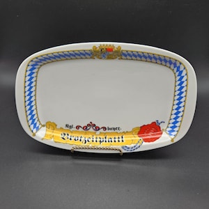 Può includere: Piatto da portata rettangolare bianco con bordo a motivo a rombi blu e bianchi. Il piatto ha un bordo dorato e presenta uno stemma e testo in tedesco. Il testo sul piatto recita "Brotzeitplattl".