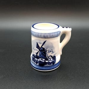 Può includere: Una piccola tazza in ceramica bianca con un design a mulino a vento blu e bianco. La tazza ha un manico e un bordo blu.