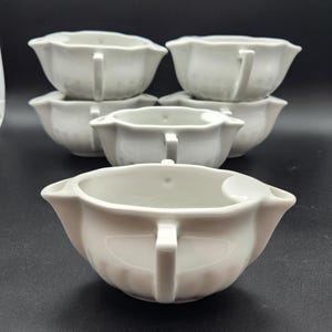 Puede incluir: Un conjunto de cuencos de cerámica blanca para salsa, cada uno con un asa y un borde festoneado. Los cuencos están dispuestos en grupo, con uno en primer plano y otros apilados detrás. Los cuencos tienen un diseño clásico y son adecuados para servir salsas o salsas.