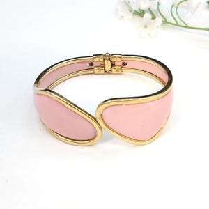May include: A hinged cuff bracelet with a gold-tone metal frame and a light pink enamel inlay. The bracelet features a clasp closure and a smooth, polished finish. The design is a modern take on a classic jewellery style.