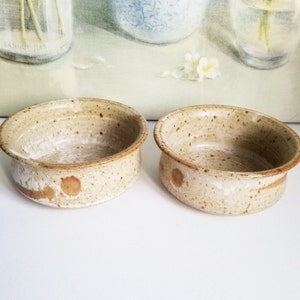 May include: Two small, speckled, ceramic bowls with a light brown glaze. The bowls have a slightly rounded shape and a white interior.