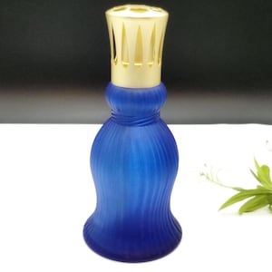 Puede incluir: Una lámpara de fragancia de vidrio azul cobalto con una tapa de metal dorado. La lámpara tiene un diseño texturizado y acanalado en el cuerpo y una tapa decorativa ranurada. La lámpara está diseñada para difundir aceites perfumados.