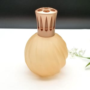 Puede incluir: Una botella decorativa de vidrio esmerilado de color melocotón con un diseño en espiral. Tiene una tapa de metal oro rosa con cortes verticales. La botella está sobre una superficie blanca.